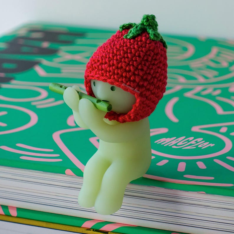 May include: A small, green, glow-in-the-dark figurine wearing a red crocheted strawberry hat plays a green flute. The figurine is sitting on a green book with pink and white markings. The book has the word "MAGIC" printed on it.