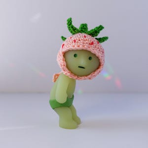 May include: A green and pink figurine wearing a crocheted strawberry hat. The figurine has a green body and a green and white striped diaper.