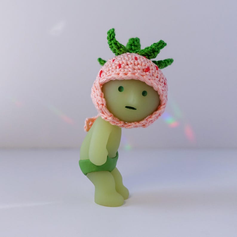 May include: A green and pink figurine wearing a crocheted strawberry hat. The figurine has a green body and a green and white striped diaper.