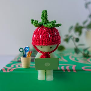 May include: A small, white figurine wearing a red crocheted strawberry hat and a scarf. The figurine is sitting on a green surface and using a laptop computer.