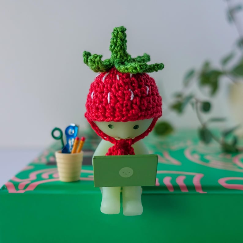 May include: A small, white figurine wearing a red crocheted strawberry hat and a scarf. The figurine is sitting on a green surface and using a laptop computer.