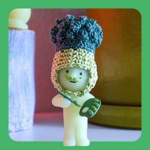 May include: A quirky figurine of an artist, featuring a light green body, a green moustache, and a crocheted hat in green and beige. The artist holds a paintbrush and a green palette. The background is blurred, with hints of other objects.