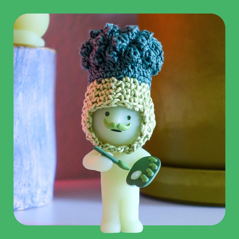 May include: A quirky figurine of an artist, featuring a light green body, a green moustache, and a crocheted hat in green and beige. The artist holds a paintbrush and a green palette. The background is blurred, with hints of other objects.