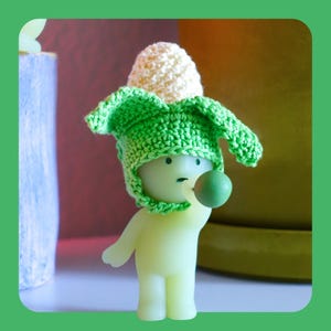 May include: A light green figurine wearing a crocheted corn-shaped hat with a white top and green leaves. The figurine is holding a small green ball. The background is blurred with shades of green and brown.