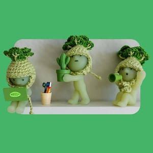 May include: Three green figurines wearing crocheted broccoli-style hats and scarves. One holds a laptop with the text "MACFLOR", another a potted cactus, and the third a green megaphone. A small desk set with scissors and pencils is also present.