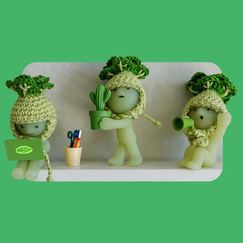 May include: Three green figurines wearing crocheted broccoli-style hats and scarves. One holds a laptop with the text "MACFLOR", another a potted cactus, and the third a green megaphone. A small desk set with scissors and pencils is also present.