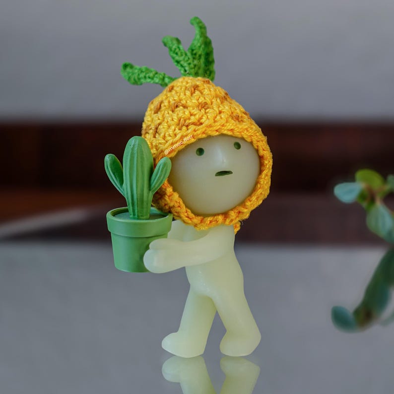 May include: A small, green glow-in-the-dark figurine wearing a yellow crocheted pineapple hat, holding a small green potted cactus.