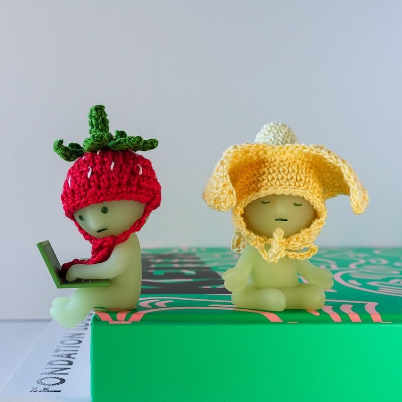 May include: Two crocheted figures, one wearing a red strawberry hat and holding a laptop, the other wearing a yellow flower hat, sitting on a green and pink patterned surface.