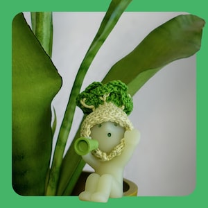 May include: A light green figurine of a person wearing a crocheted hat and holding a green object. The figurine is set against a backdrop of green plant leaves, creating a playful and natural scene.