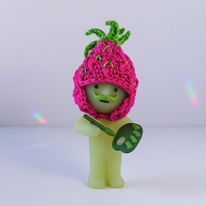 May include: A green and pink crocheted strawberry hat with a green stem on top. The hat is worn by a green glow-in-the-dark figurine with a mustache. The figurine is holding a green paint palette and a paintbrush.