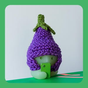 May include: A light green figurine of an aubergine wearing a crocheted purple hat with green leaves. The figurine is holding a small green rectangle, resembling a phone. The background is white.