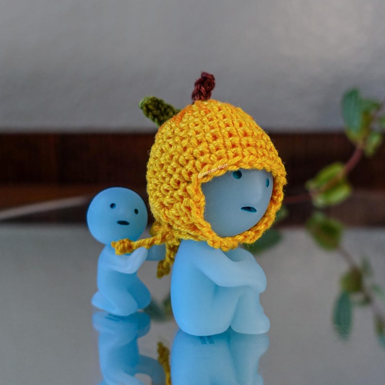 May include: Two blue figurines, one wearing a yellow crocheted hat with a brown stem, are sitting on a reflective surface. The figurine in the hat is sitting with its legs crossed and its arms folded. The other figurine is standing and holding onto the hat's stem.