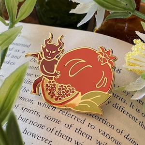 May include: A gold-colored enamel pin featuring a red pomegranate and a small devil figure. The pin is set against a backdrop of a book page with text and surrounded by greenery and white flowers.