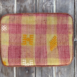 May include: Rectangular cushion with a woven, plaid pattern in shades of red and yellow. The cushion features embroidered orange geometric designs. The edges are finished with a brown border. This decorative cushion is suitable for home decor.