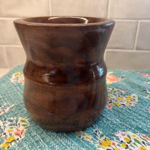 May include: A dark brown wooden vase with a smooth, polished finish. The vase has a wide mouth and a narrow base, giving it a unique shape.
