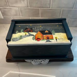 May include: A rectangular wooden box with a dark blue finish and a sliding lid. The lid features a hand-painted winter scene with two orange houses, a small snowman, yellow stars, and green pine trees against a dark blue night sky background.