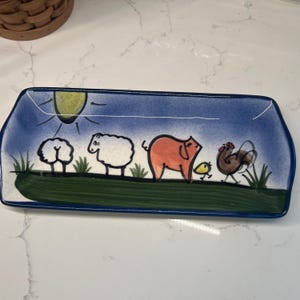 May include: A blue and white ceramic rectangular serving dish with a green rim. The dish features a whimsical painted scene of a pig, two sheep, a rooster, and a chick in a field with a yellow sun.
