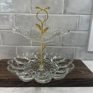 May include: A two-tier serving stand featuring clear pressed glass plates with scalloped edges and a textured pattern. The central support rod is made of gold-toned metal and is shaped like a slender stem with decorative leaves and a loop handle.