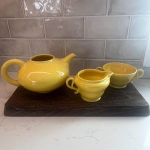 May include: A yellow ceramic tea set, including a teapot, creamer, and sugar bowl, sits on a dark wooden tray. The teapot has a rounded body and a curved spout. The creamer and sugar bowl have handles.