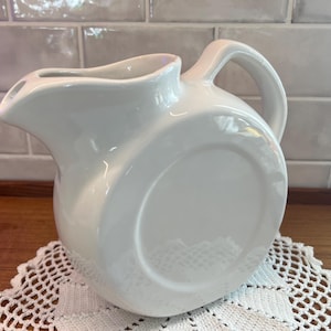 May include: A white ceramic pitcher with a curved spout and a large handle. The pitcher has a rounded body and sits on a white lace doily. The background features a light-colored tiled wall and a wooden surface.