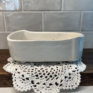 May include: A white ceramic bathtub-shaped planter with a smooth, glossy finish and four small feet. The vessel features an elongated, rounded rectangular basin with slightly uneven, hand-crafted edges and a clean, minimalist white surface.