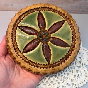 May include: A round, ceramic trivet with a floral design. The center features a green background with a maroon flower design. The outer edge is a light brown with a textured, scalloped edge. The trivet is approximately 6 inches in diameter.