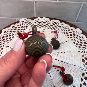 May include: Three small, antique bronze bells with a spiral design and red cords. The bells are held against a white lace doily on a wooden tray. The bells have a loop at the top for hanging.