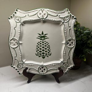 May include: White square decorative plate with a green pineapple design in the center. The plate has a decorative border with green accents and is displayed on a wooden stand. The plate is approximately 12 inches square.