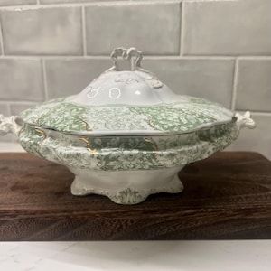 May include: A white and green ceramic serving dish with a lid. The dish features a floral pattern in green and gold accents. The dish is on a dark brown wooden surface.