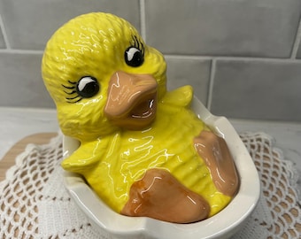 May include: A ceramic yellow duckling figurine emerging from a white eggshell. The duckling has black eyes, an orange beak, and orange feet. The eggshell has a scalloped edge. A decorative item.