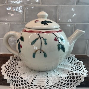 May include: A cream-colored ceramic teapot featuring a hand-painted design of green holly leaves, red berries, and a thin red ribbon encircling the upper body. The teapot includes a matching lid with a knob handle and a curved spout.
