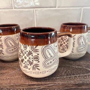 May include: Three ceramic mugs with a brown rim and handle, featuring a cream-colored body. Each mug is decorated with a brown paisley design and a series of small square and star-like shapes. The mugs are on a wooden surface.
