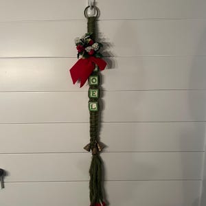 May include: A vertical wall hanging featuring a green macrame cord topped with a metal ring. It includes a red fabric bow, artificial holly berries, and four square blocks spelling NOEL in green letters. The base has small metal bells and a tassel.