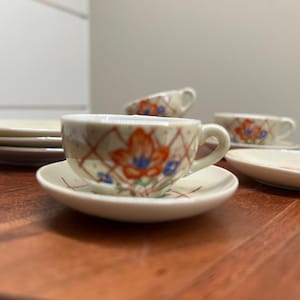 May include: A set of miniature teacups and saucers with floral designs. The cups and saucers are white with a delicate floral pattern in shades of orange, blue, and green. The cups have a small handle and the saucers are round.