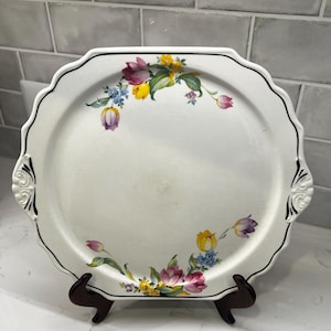 May include: A white, scalloped-edge serving platter with a black trim and floral design. The flowers are in shades of pink, yellow, and purple, with green leaves. The platter has decorative handles and is displayed on a wooden stand.