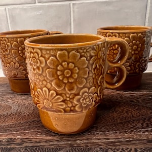 May include: Three brown ceramic mugs with a floral pattern. The mugs have a unique handle design and are placed on a wooden cutting board. The mugs are a warm brown color and have a glossy finish. The floral pattern is embossed on the surface of the mugs.