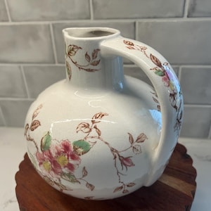 May include: A white ceramic pitcher with a floral design. The pitcher has a round body and a handle. The flowers are pink, brown, and green.