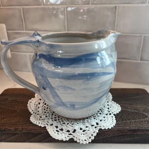 May include: A handmade ceramic pitcher with a blue and white swirl design. The pitcher has a curved handle with a decorative finial and a spout for easy pouring. It sits on a white crocheted doily.