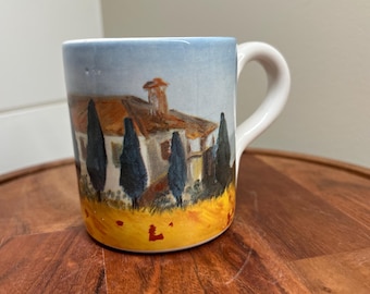 May include: White ceramic mug with a painted scene of a house with a red roof and trees in a field. The mug has a white handle.