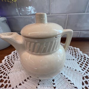 May include: A cream-colored ceramic teapot with a lid and handle. The teapot has a spout and is sitting on a white, lacy doily. The background features a tiled wall and a wooden surface.