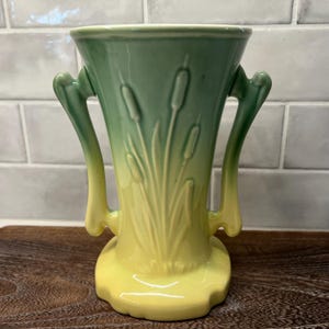 May include: A two-handled ceramic vase with a flared top and a round base. The vase is painted in shades of green and yellow, with a cattail design. The handles curve outward from the body of the vase.