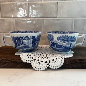 May include: Two white ceramic teacups featuring a blue transferware pattern depicting detailed landscape scenes with architectural ruins and trees. Each cup has a wide rim, a footed base, and a distinct angular handle with a blue floral border.