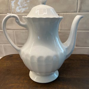 May include: A white ceramic teapot with a lid, handle, and spout. The teapot has a fluted body and sits on a round base. The background is a light-colored tiled wall and a wooden surface.