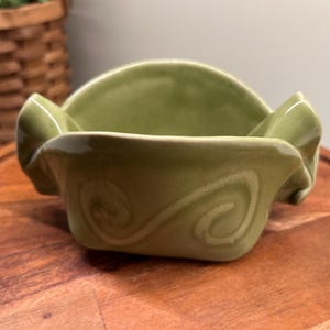 May include: A green ceramic bowl with a wavy, ruffled rim and a swirl design. The bowl is sitting on a wooden surface.