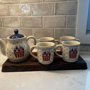 May include: A ceramic tea set featuring a teapot and three mugs, all decorated with a fruit basket design. The teapot has a blue lid and handle, while the mugs have a matching pattern. The set is displayed on a dark wooden tray.