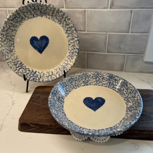 May include: Two ceramic plates with a blue heart design. The plates have a cream-colored center and a blue speckled border. One plate is displayed on a black metal stand. The other plate is on a wooden board.
