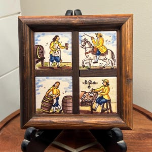 May include: A framed set of four ceramic tiles depicting scenes of people working in a winery. The tiles are painted in a rustic style with brown, yellow, blue, and white colors. The tiles are set in a wooden frame with a dark brown finish.