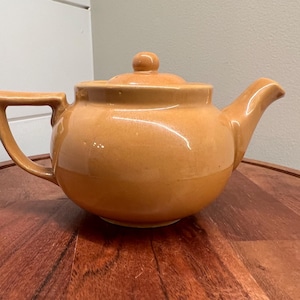 May include: A ceramic teapot in a warm, light brown color. The teapot has a rounded body, a curved spout, and a handle. A lid with a small knob sits on top. The teapot is placed on a wooden surface.