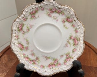 May include: A white porcelain saucer with a scalloped edge, adorned with pink roses and green leaves. The rim is trimmed with gold. The saucer is displayed on a black stand, showcasing its delicate floral design.