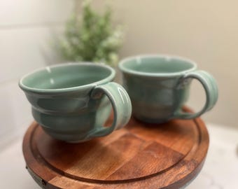 May include: Two light blue ceramic mugs with a textured, wavy design. The mugs have a rounded shape and a handle on the side. The mugs are sitting on a wooden surface.